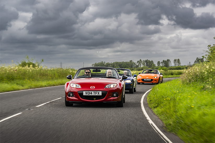 A red, blue, and orange Mazda MX-5 driving along a road with tall grass on either side