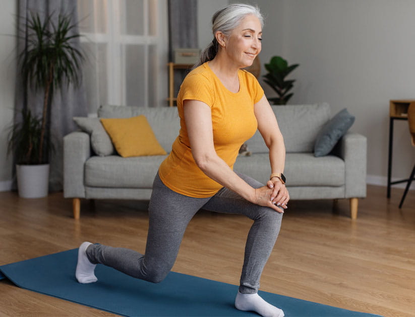 Mature woman doing demonstrating standing hip flexor lunges on yoga mat in living room
