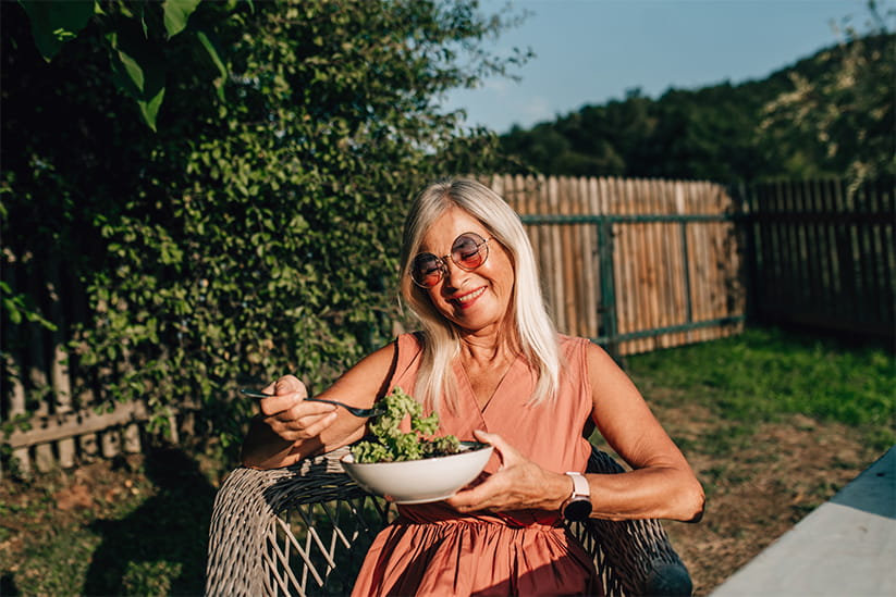 Senior woman is sitting in the garden, eating salad and smiling
