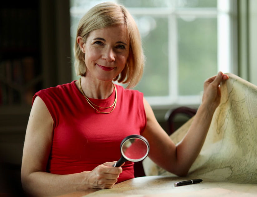 Lucy Worsley sitting at a table with a magnifying glass in one hand and holding up the corner of an old map with the other