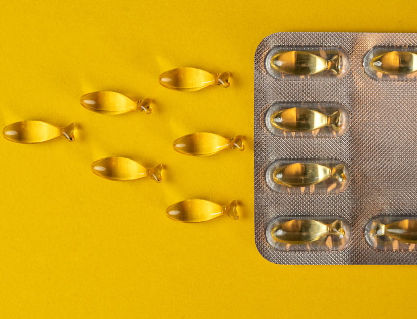 Close-up of a blister pack of Omega-3 or vitamin D capsules in the shape of a fish on a yellow background, surrounded by scattered capsules 