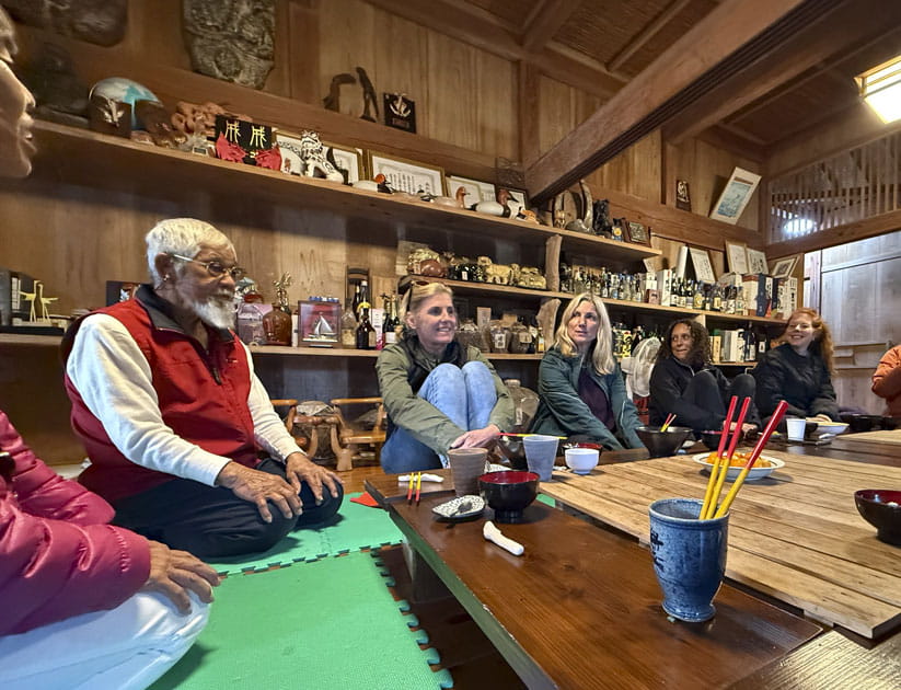 A group of people kneeling on mats around a low wooden table with bowls and chopsticks on the surface.