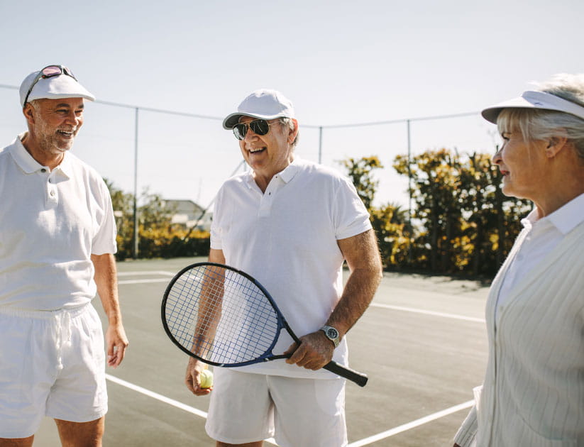 Group of three senior people playing tennis on a sunny day - standing on tennis court holding rackets and balls.