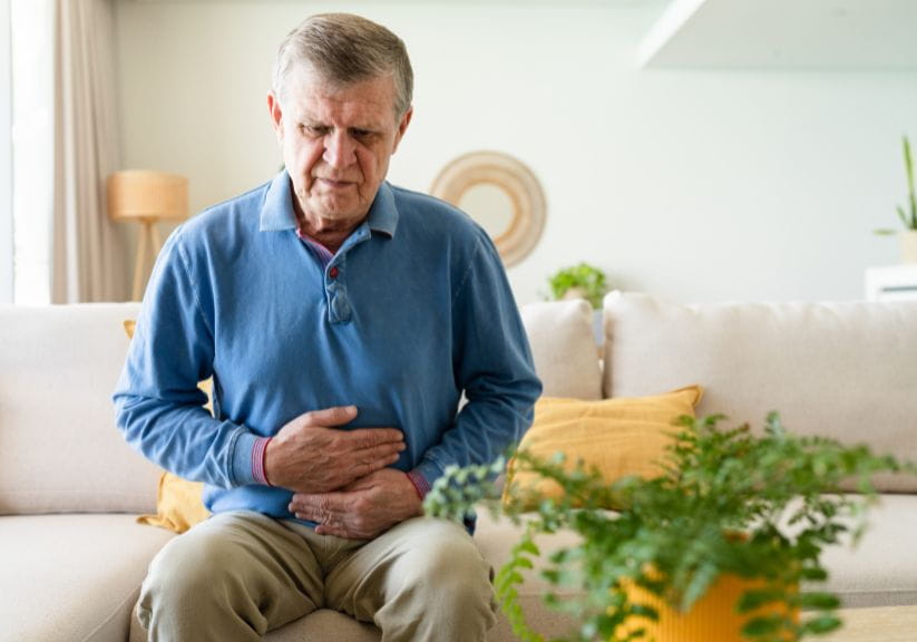 A man in a blue jumper clutches his stomach in pain, while stitting on a sofa