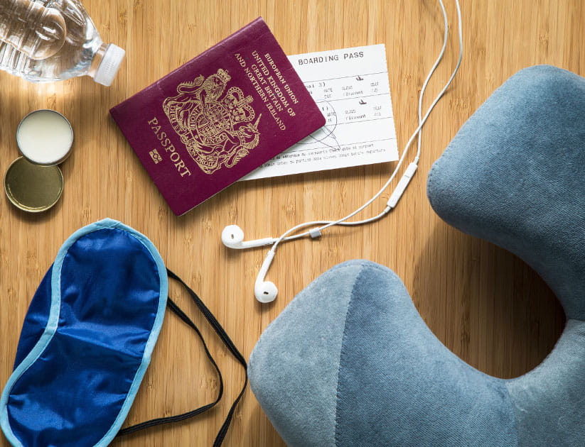 Overhead view of items needed for aeroplane travel including a neck pillow, headphones, lip balm, eye mask and a bottle of water