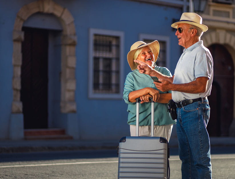 Happy senior tourist couple wearing hats outside in the sunshine with a suitcase