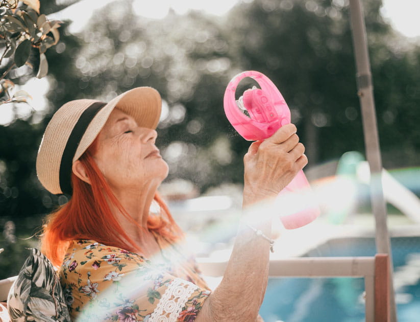 Senior woman in floral summer dress enjoys cool air from electric plastic pink fan on summer afternoon 