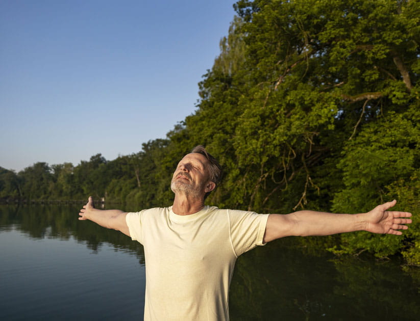 Mature man with arms outstretched and eyes closed facing up to the sunlight near a body of water
