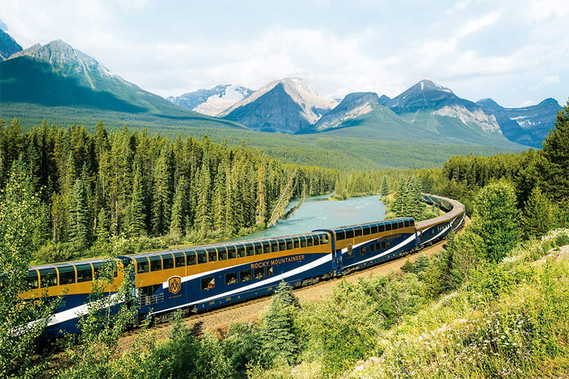 The Rocky Mountaineer train alongside a river with mountains in the background