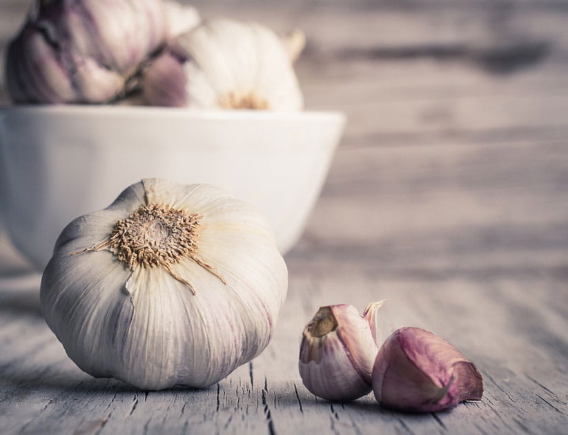 A garlic bulb with two cloves on a wooden surface with a with bowl filled with more garlic bulbs in the background