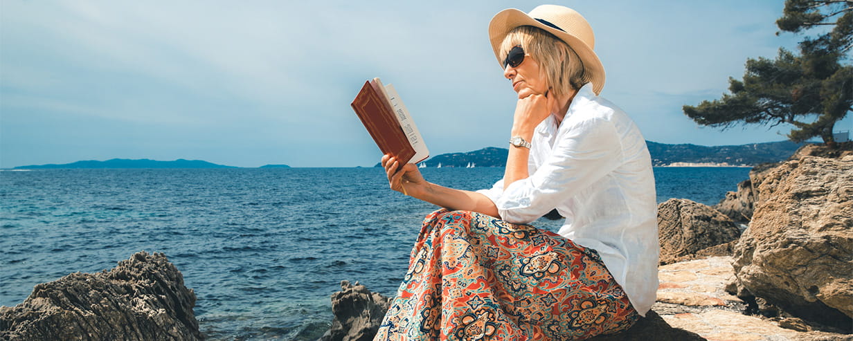 Senior woman reading a book on a rocky seashore in Provence, France.