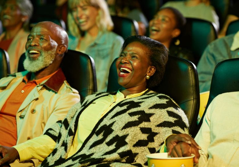 A middle aged black couple sit in the cinema laughing while looking at the screen