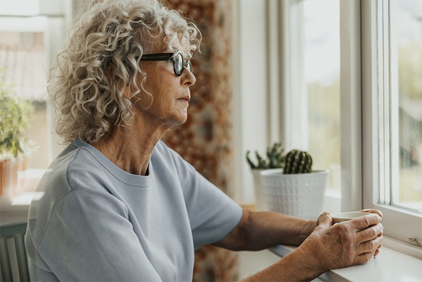 Thoughtful senior woman holding coffee cup while looking through window at home