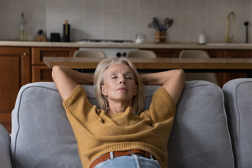 Calm peaceful elder lady relaxing with closed eyes on comfortable sofa at home, taking deep breath of fresh air, thinking, dreaming, reducing stress, practicing meditation, mental exercises
