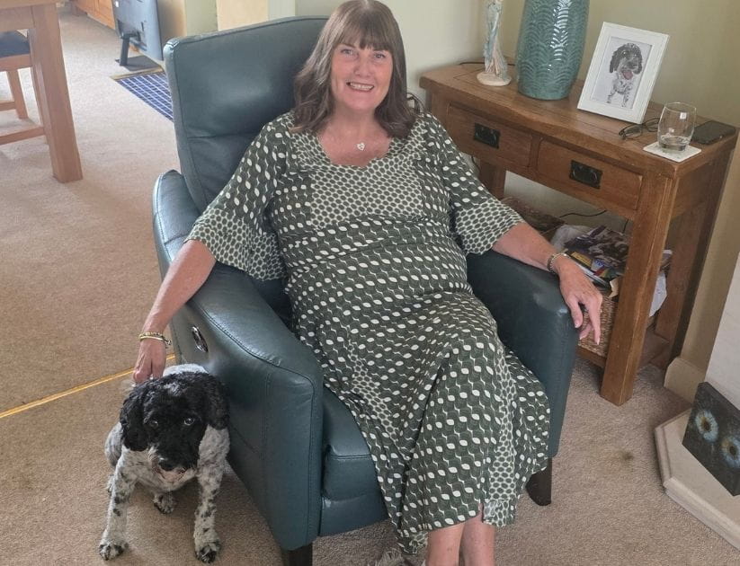 A photograph of Sue on a chair in her living room with her dog beside her