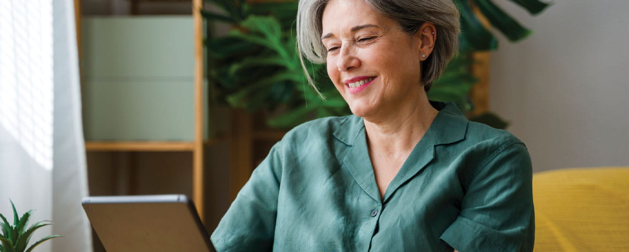 Mature smiling woman in a green blouse reading tablet indoors in casual home setting