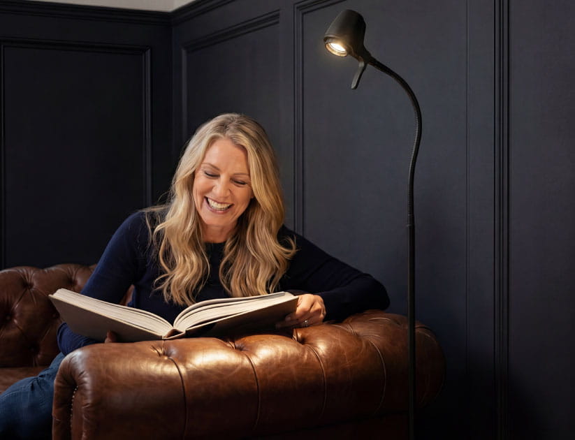 Mature smiling woman on a brown leather sofa reading a book illuminated by a Serious Readers lamp