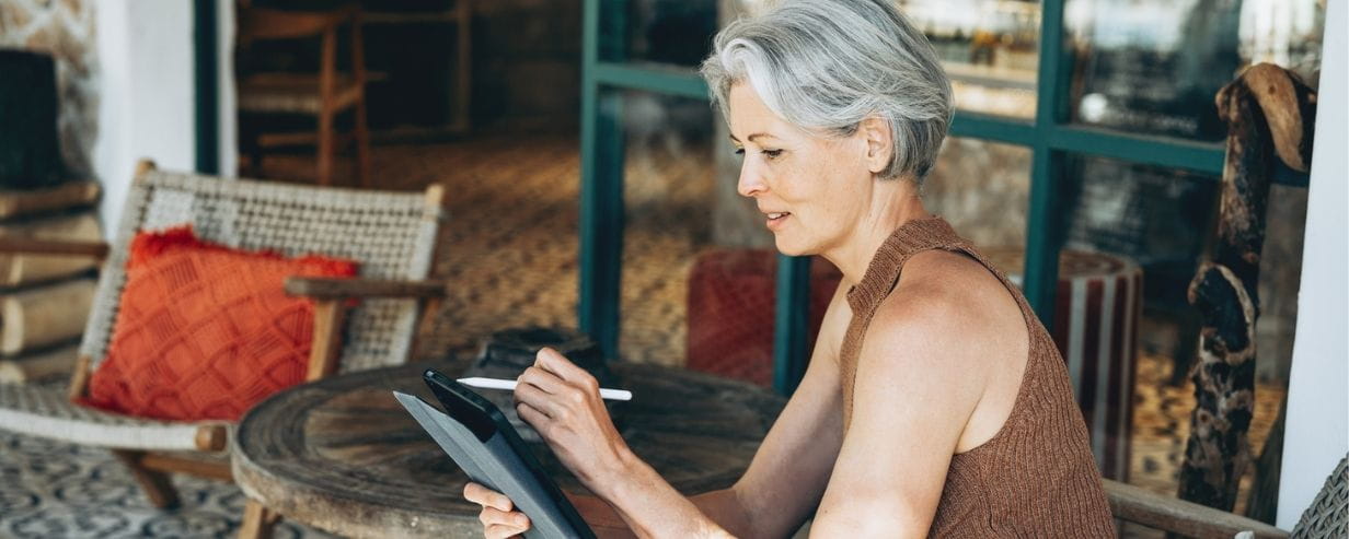 Mature woman with short gray hair sits comfortably on a patio chair using a digital tablet and stylus