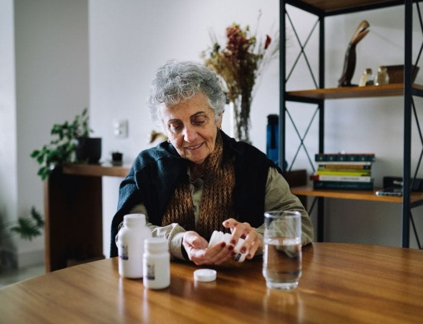 Woman sitting at a table, pouring pills from a bottle into her hand, with a glass of water nearby