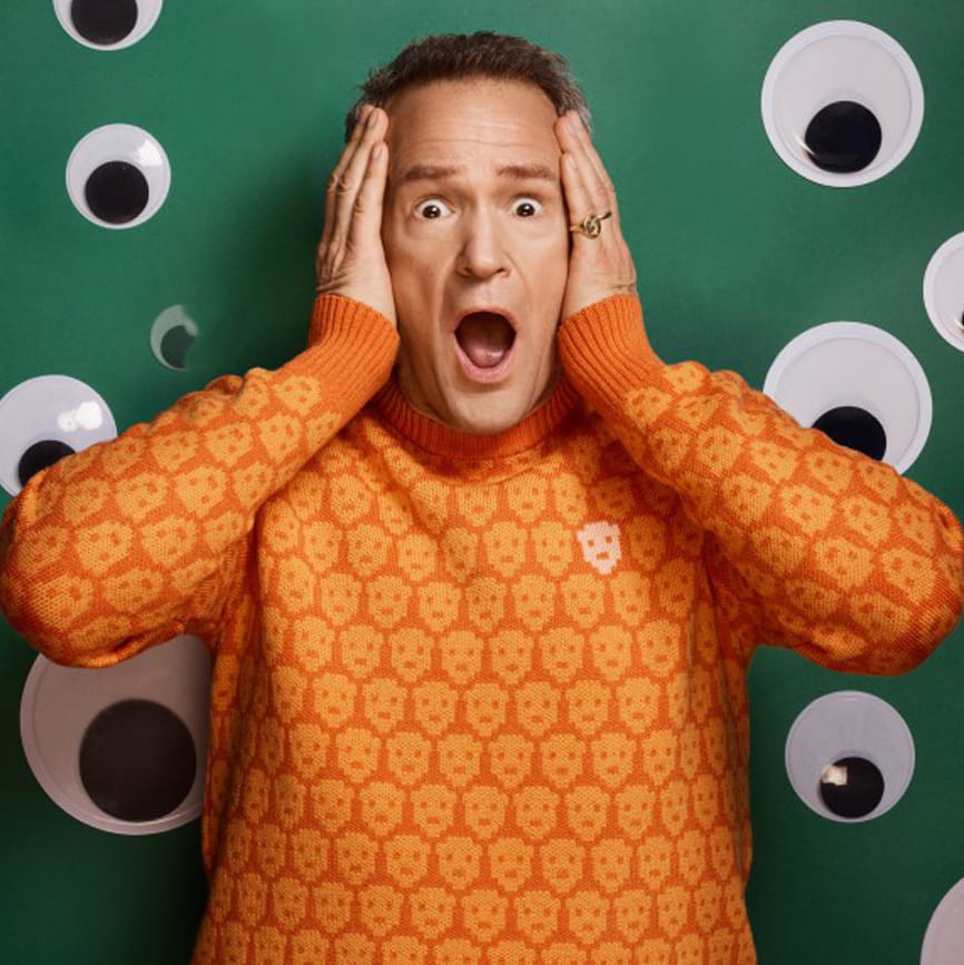 Alexander Armstrong in an orange jumper with a green background covered in googly eyes