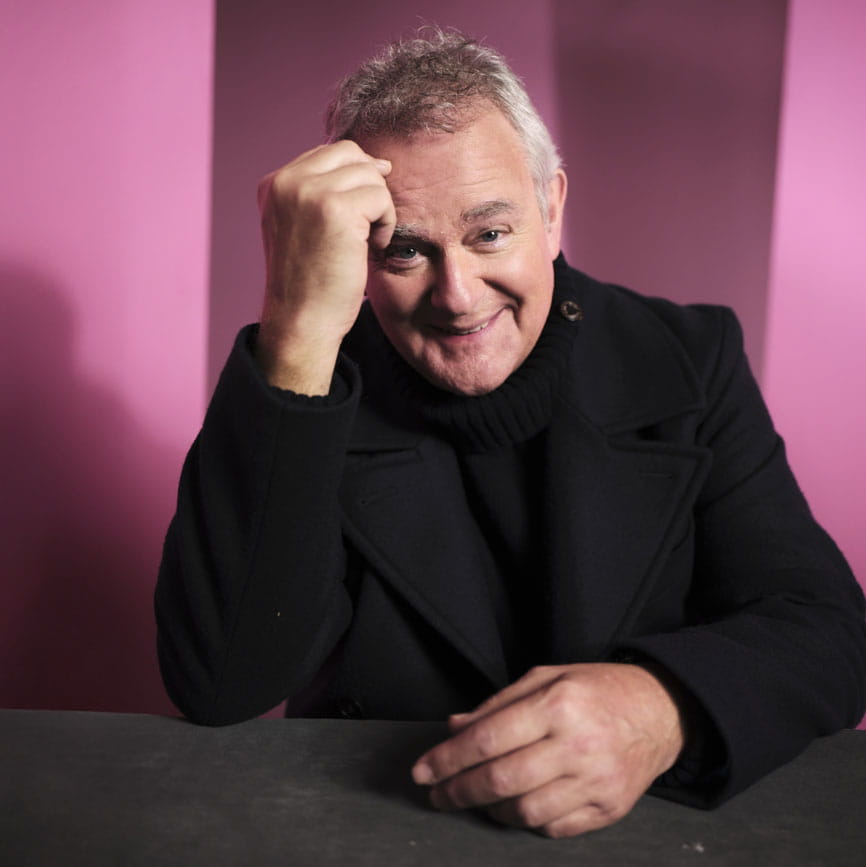 Head and shoulders photo of Hugh Bonneville in a black coat and roll-neck jumper with a pink background