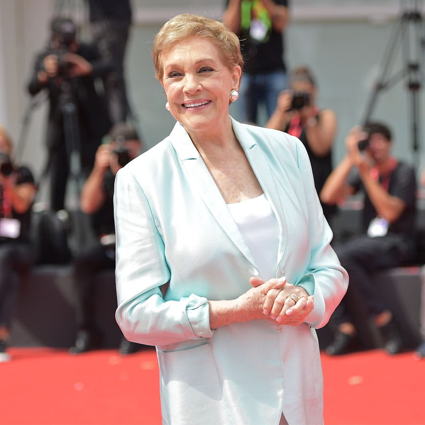 Julie Andrews on a red carpet with people holding cameras blurred in the background