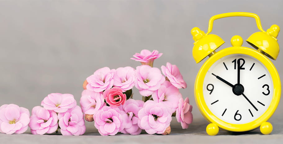 A bright yellow analogue alarm clock next to some flowers