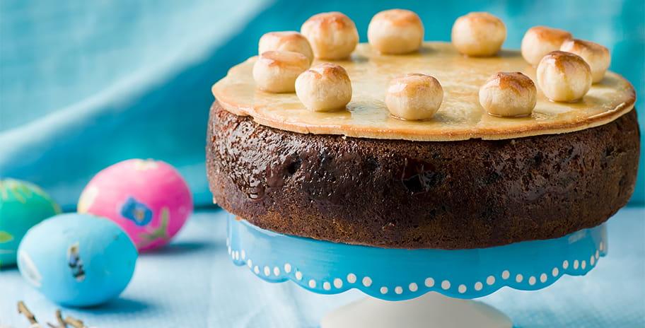 An Easter Simnel cake on a blue background, with some colourful eggs in the corner