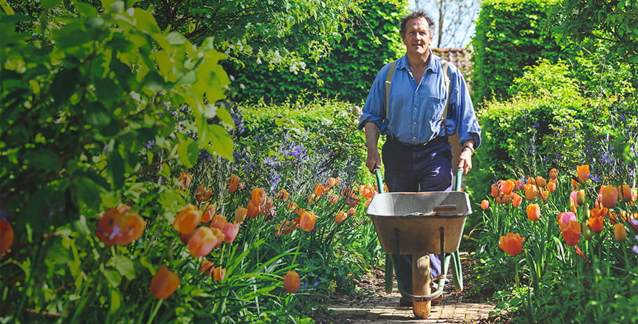 Monty Don pushing a wheelbarrow along a garden path surrounded by flowers
