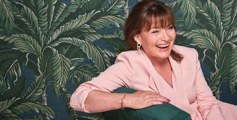 Lorraine Kelly in a pink suit sitting on a sofa with a green palm tree wallpaper behind her