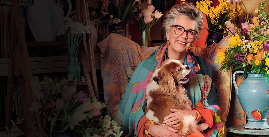 Prue Leith sitting in an armchair while holding a dog and smiling