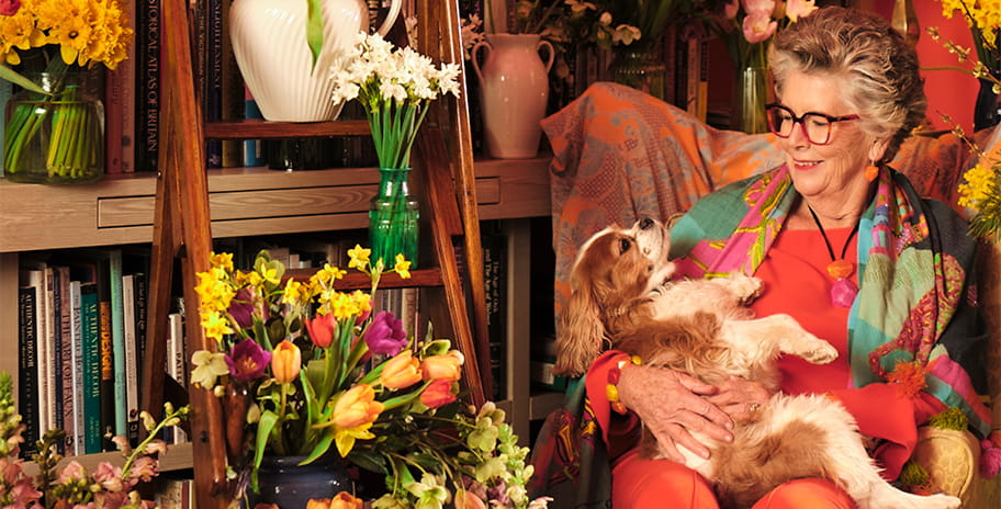 Prue Leith sitting in an armchair while holding a dog and smiling
