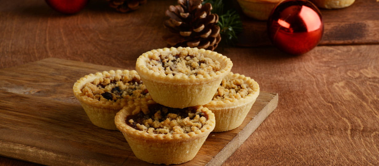 A stack of crumble mince pies on a wooden table with Christmas decorations visible in the background