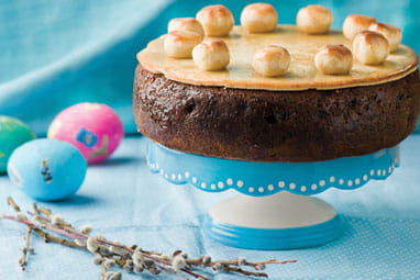 A traditional Easter Simnel cake on a stand with a blue frill with painted Easter eggs on the right