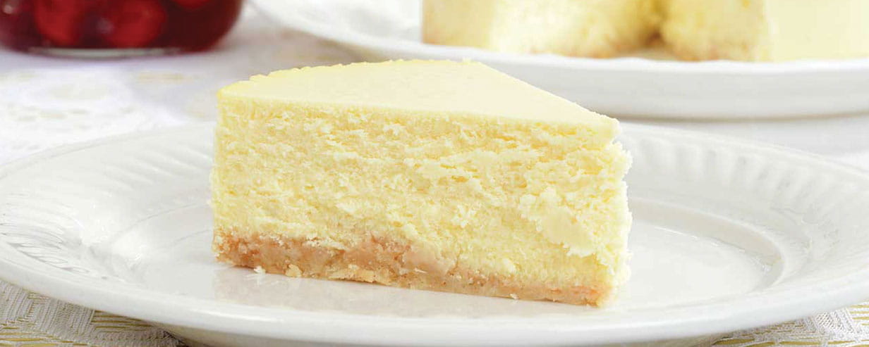 A slice of New York cheese cake with the rest of the cake in the background and a bowl of berries.