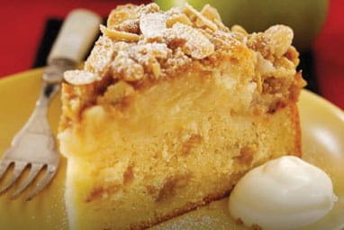 Close up of a slice of German apple cake, topped with flaked almonds, on a plate with a fork and a dollop of cream