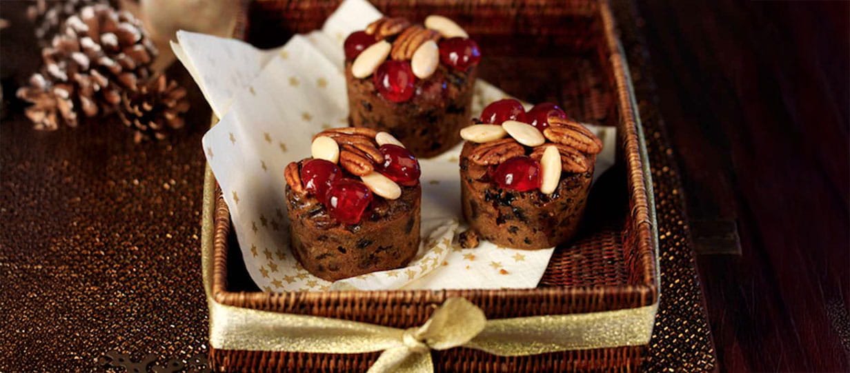 Three mini Christmas cake muffins topped with glace cherries and pecan nuts in a square basket lined with gold star paper