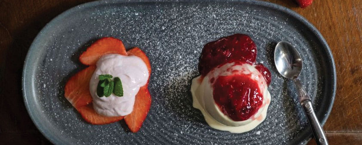 A grey plate with sliced strawberries and panna cotta on top and next to it panna cotta with red berry sauce