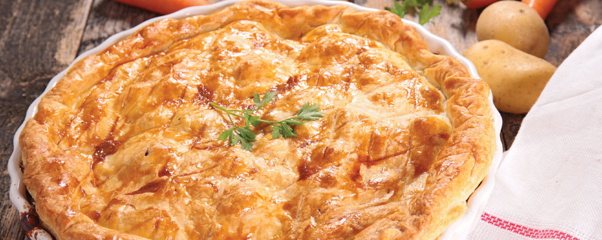 A golden crusted pie in a white dish with potatoes and carrots in the background
