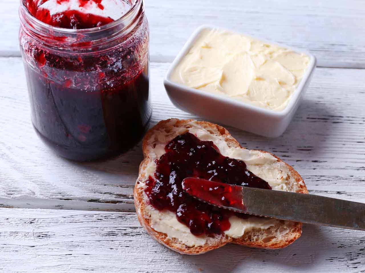 Fresh bread being spread with jam, on light wooden background