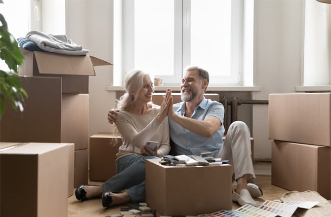 Two older people sitting on the floor high fiving surrounded by packing boxes