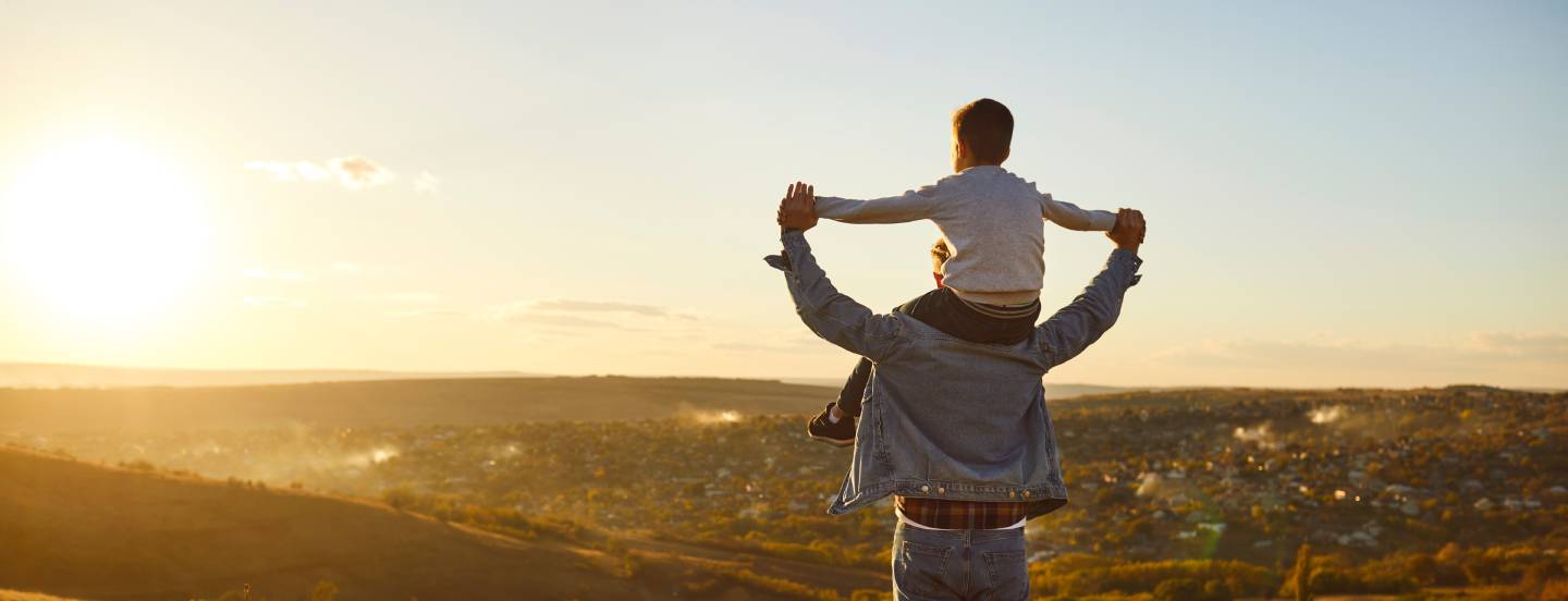 ack view of a little child boy sitting on his fathers shoulders holding hands and looking into the distance enjoying sunset.