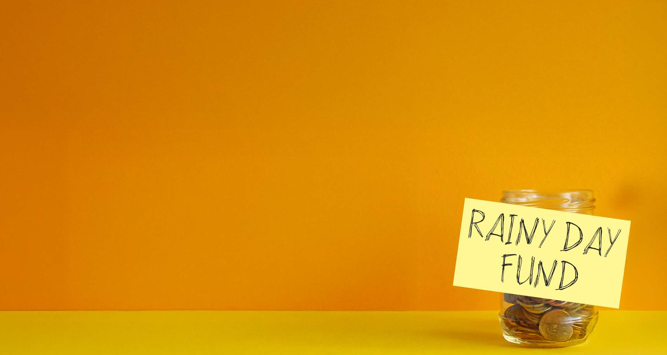 A glass jar with coins in with a sticky note that reads "rainy day fund"