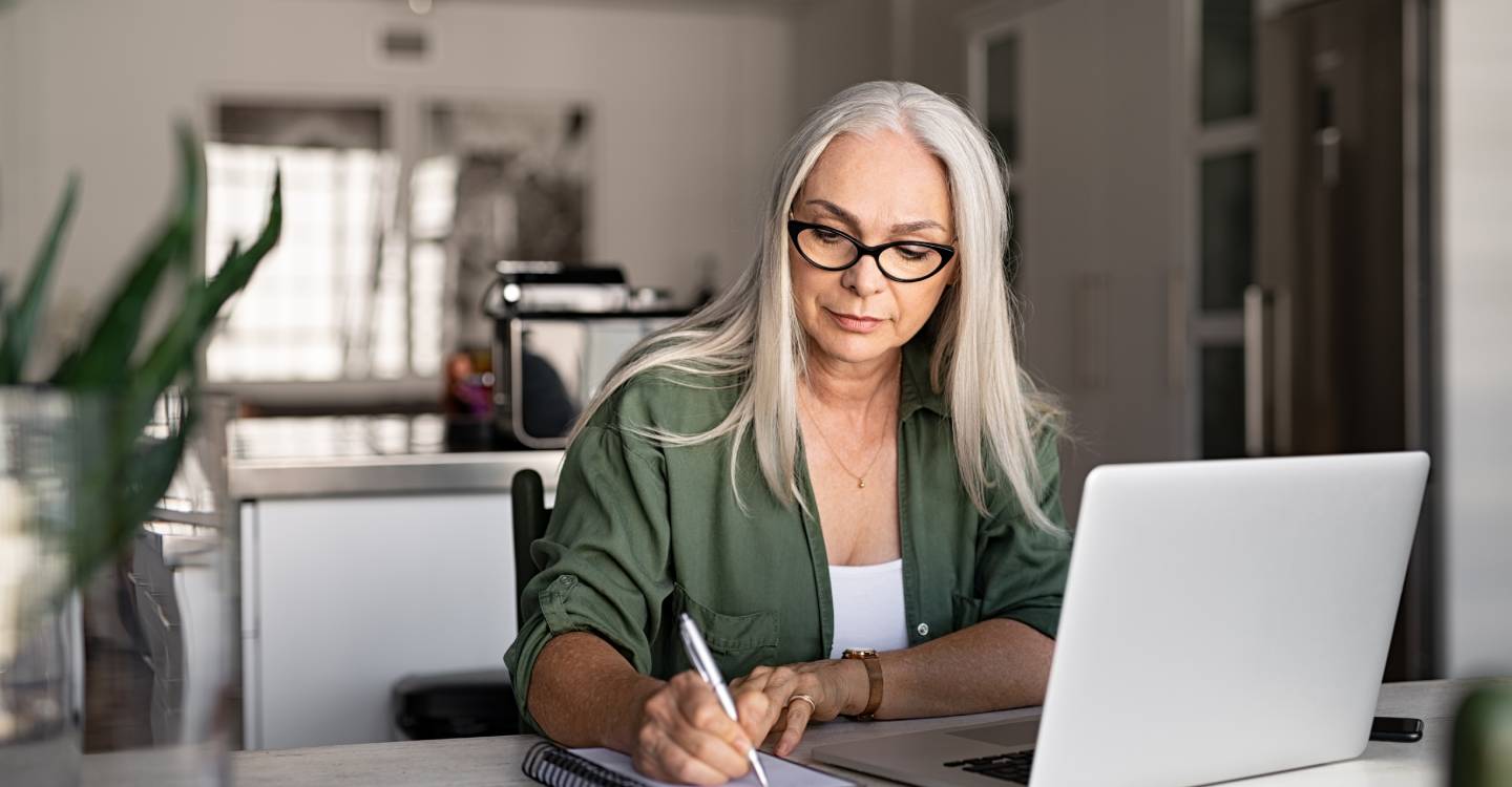 Senior woman taking notes in notebook while using her laptop at home
