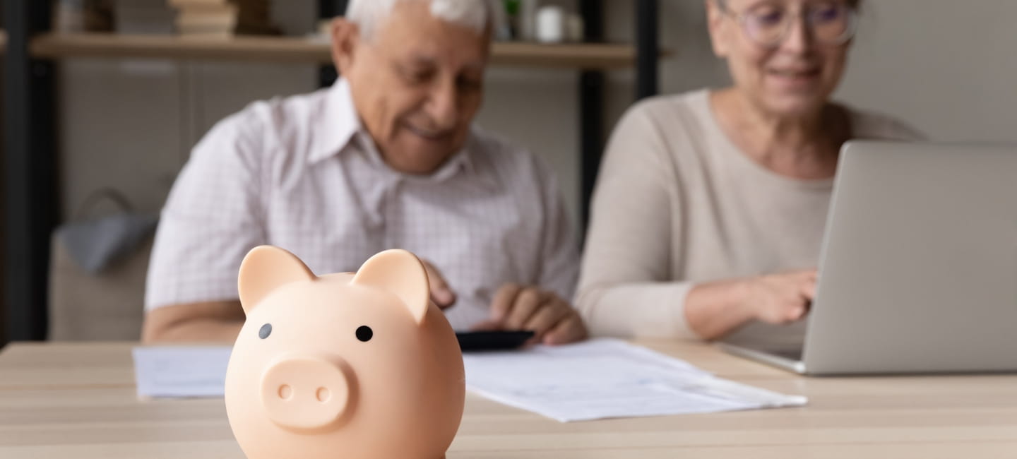 Focus on piggybank on table, on background frugal elderly couple make payment using e-bank on laptop, calculates earning smile feel happy. Family budget control, management, investment, profit concept.