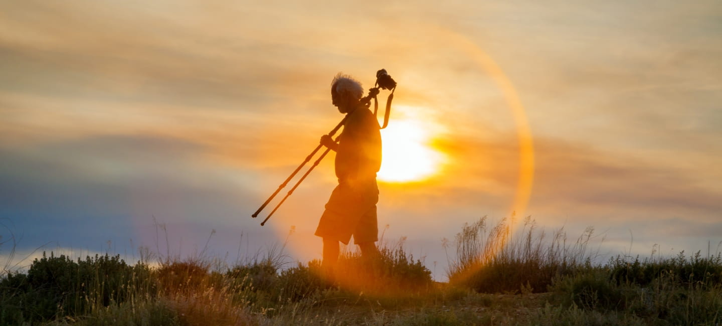 A seasoned photographer with grey hair blowing in the wind, walks at sunset with his tripod resting on his shoulder.  He is in partial silhouette with the sun directly behind him.