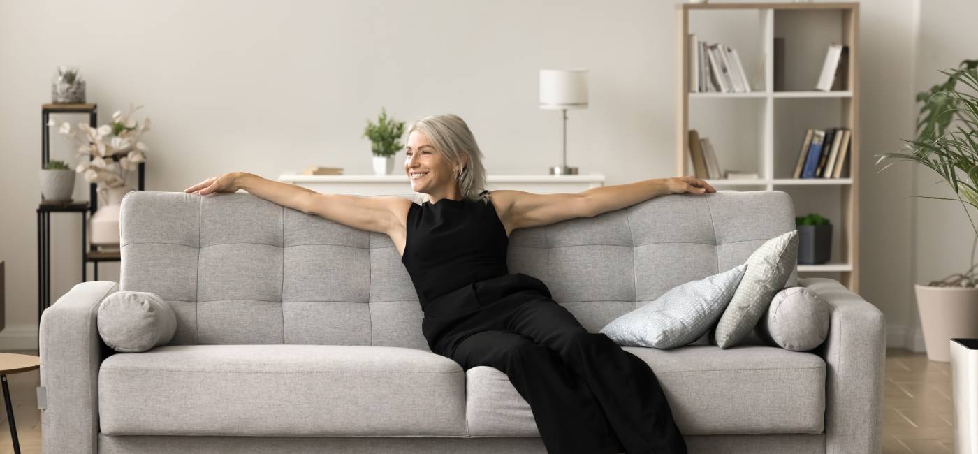 Happy thoughtful mature blonde woman relaxing on home sofa, leaning on soft back with open hands, taking deep breath, enjoying fresh pure air, domestic comfort, looking away, thinking, dreaming