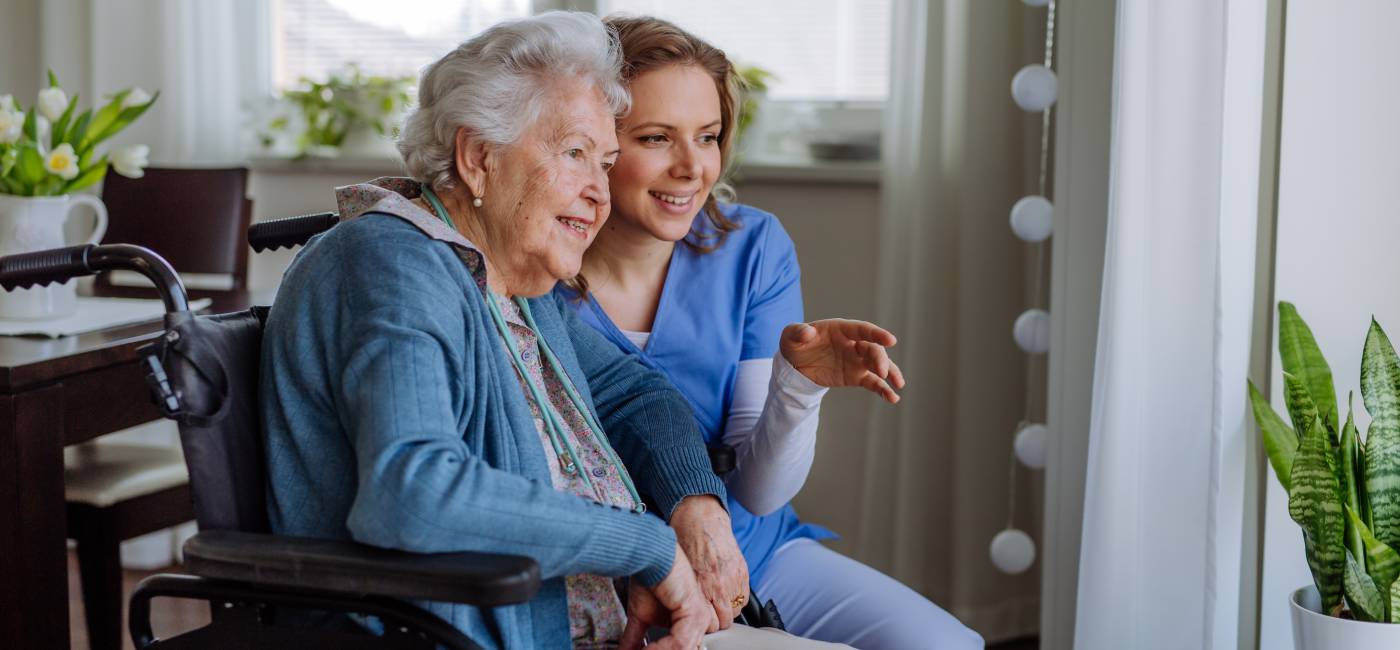 "An elderly woman smiling while sitting in a wheel chair with a nurse sitting next to her pointing out of a window"