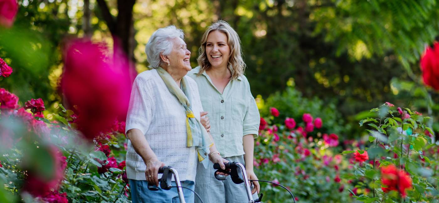 An elderly woman with a walking aid smiles while walking with her granddaughter in a flower-filled field.
