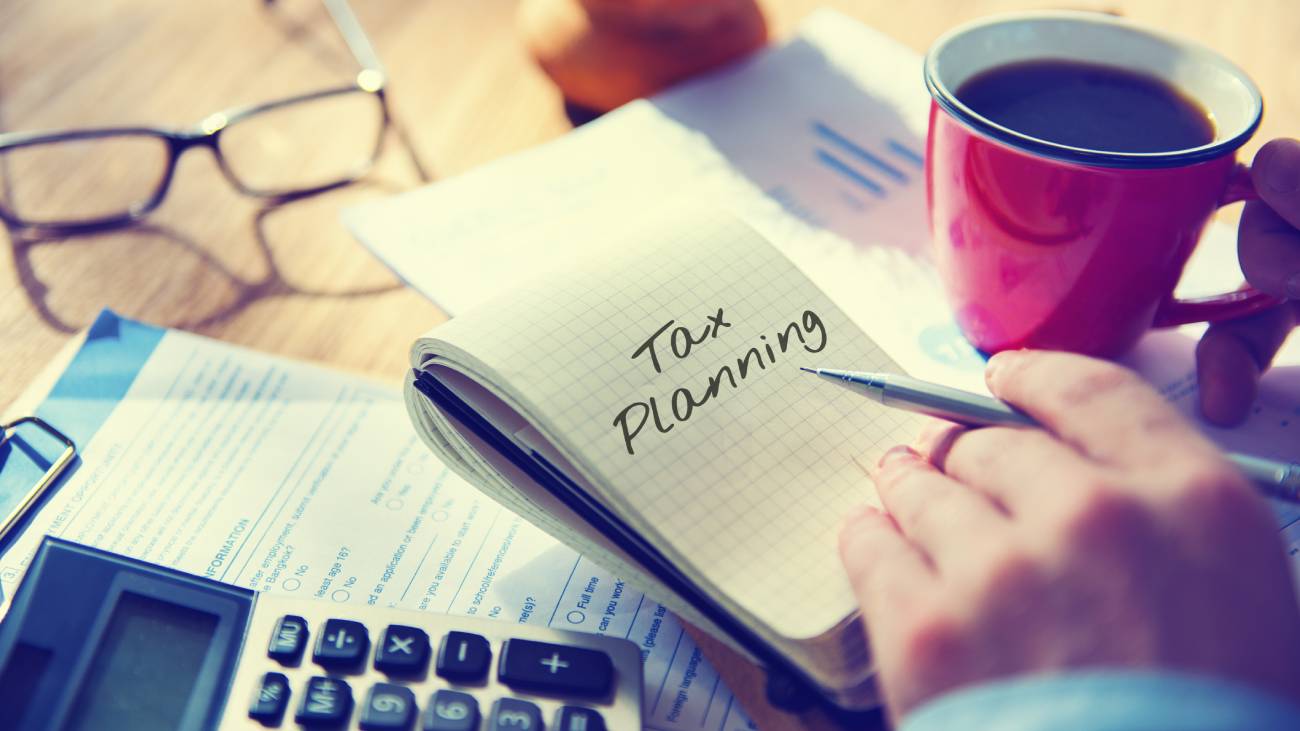 A person writing tax planning on a note pad working out taxes with a calculator as they hold a pen